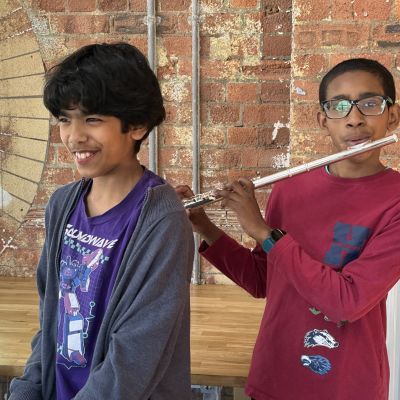 Two young boys, the one on the left is smiling and playing the keyboard, the one on the right is playing the flute, they are in a well lit room with red brick wall